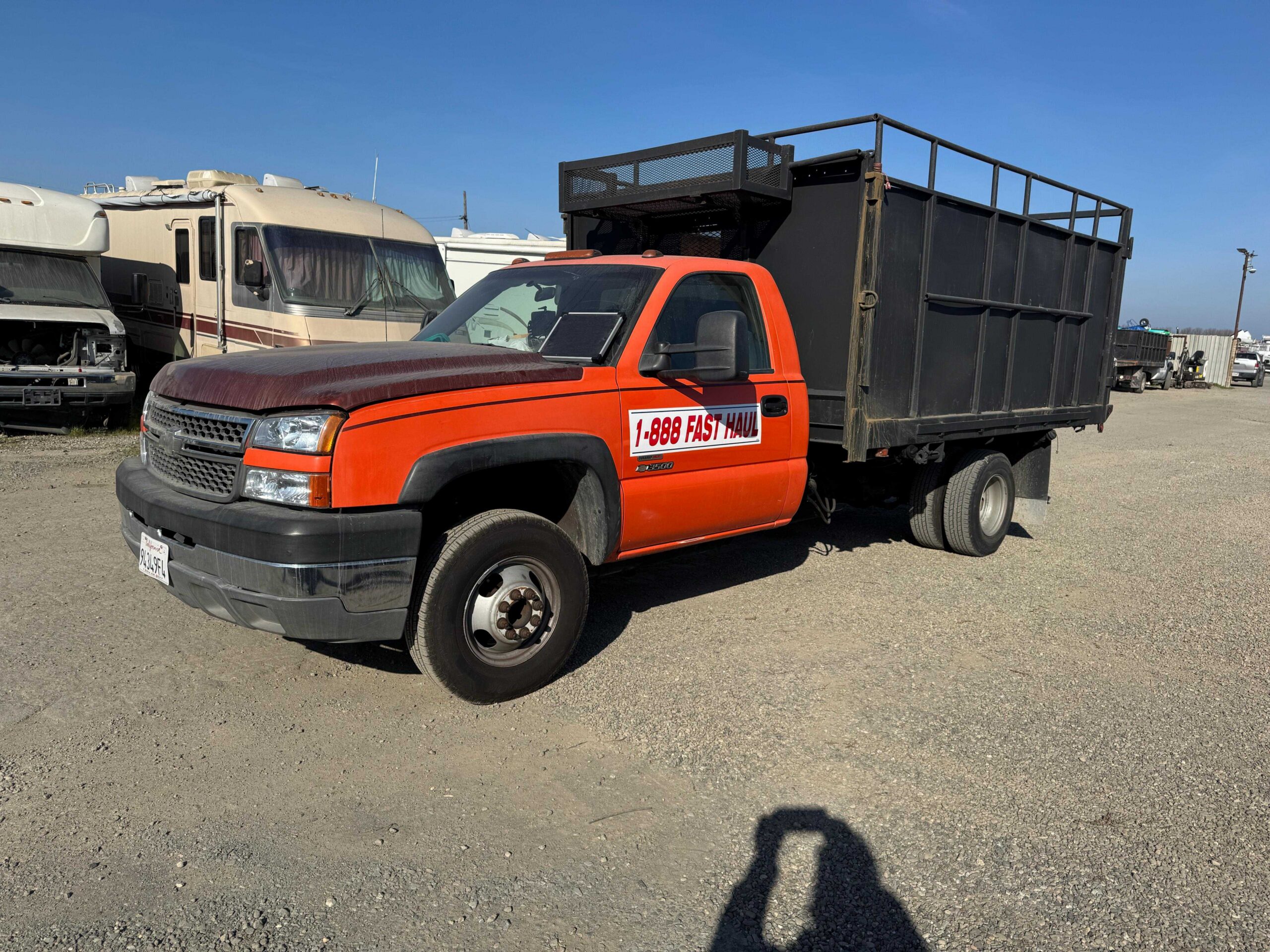 High-Capacity Hauling Fleet Orange flatbed utility dump truck with high-sided black steel box parked on gravel under a blue sky.