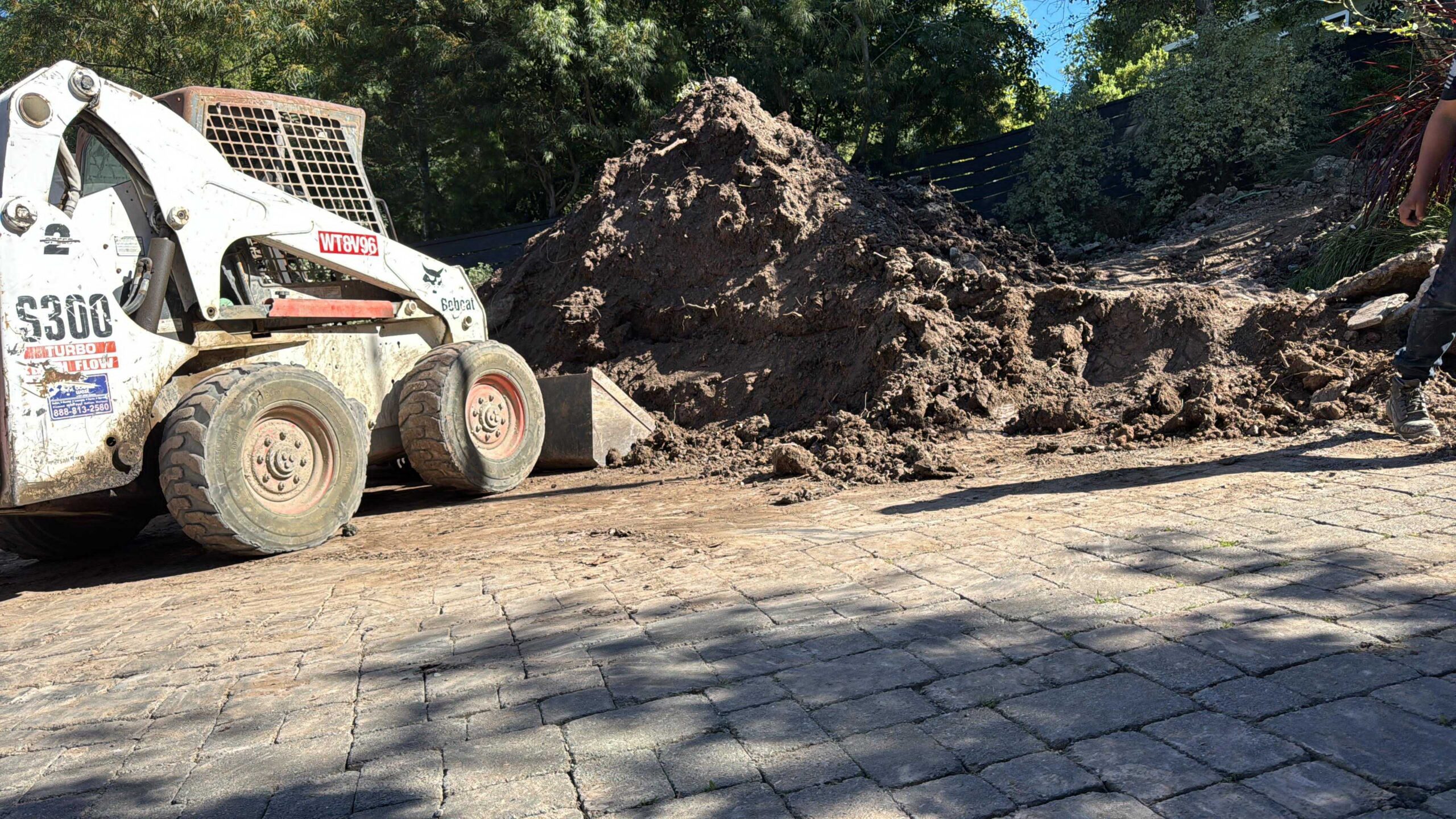Precision Bobcat Excavation Work White Bobcat skid steer loader excavating a massive pile of soil on a residential paver stone driveway.
