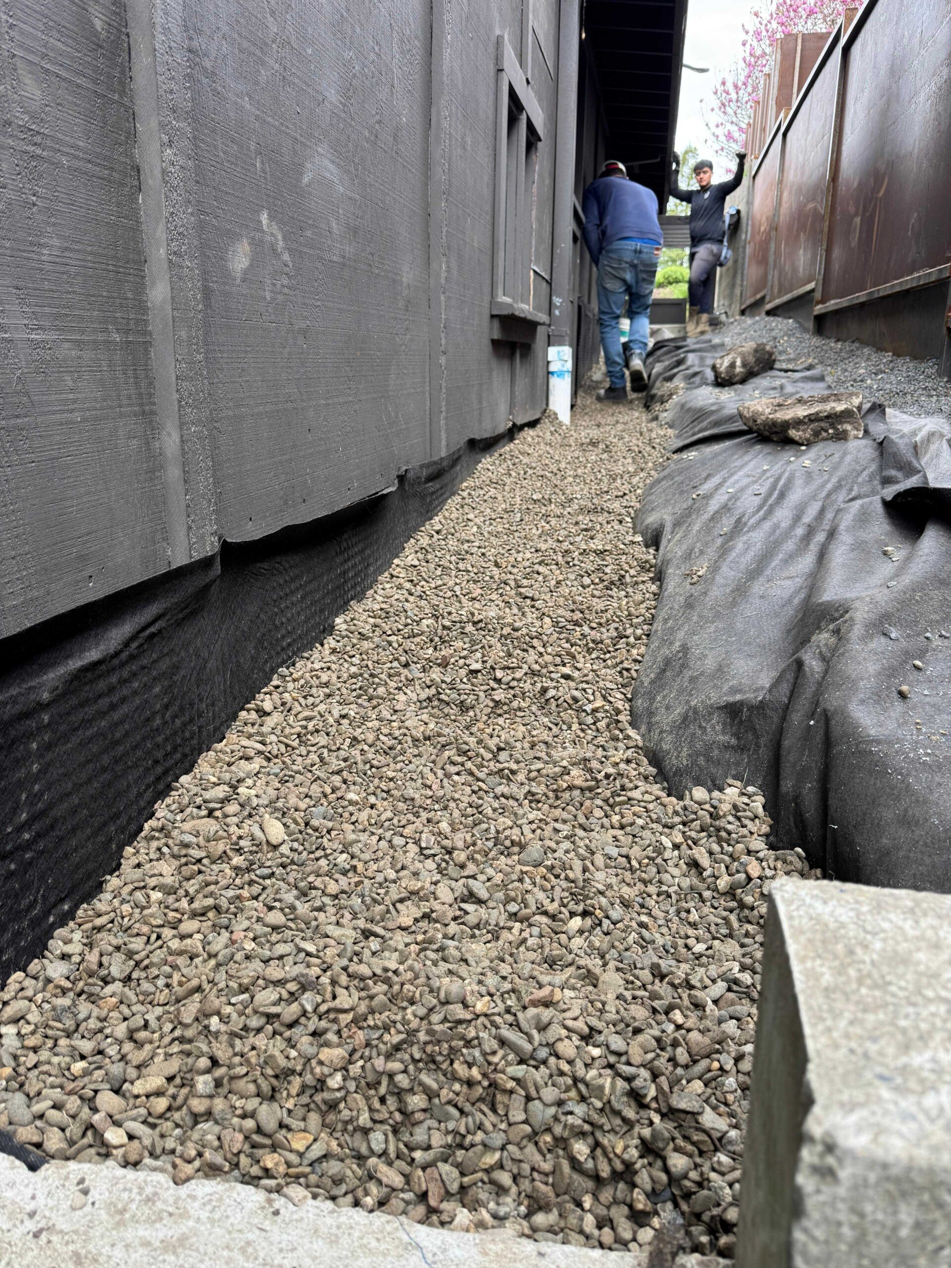 Residential Trenching and Drainage Solutions Construction workers backfilling a backyard drainage trench with gravel along the foundation of a house.
