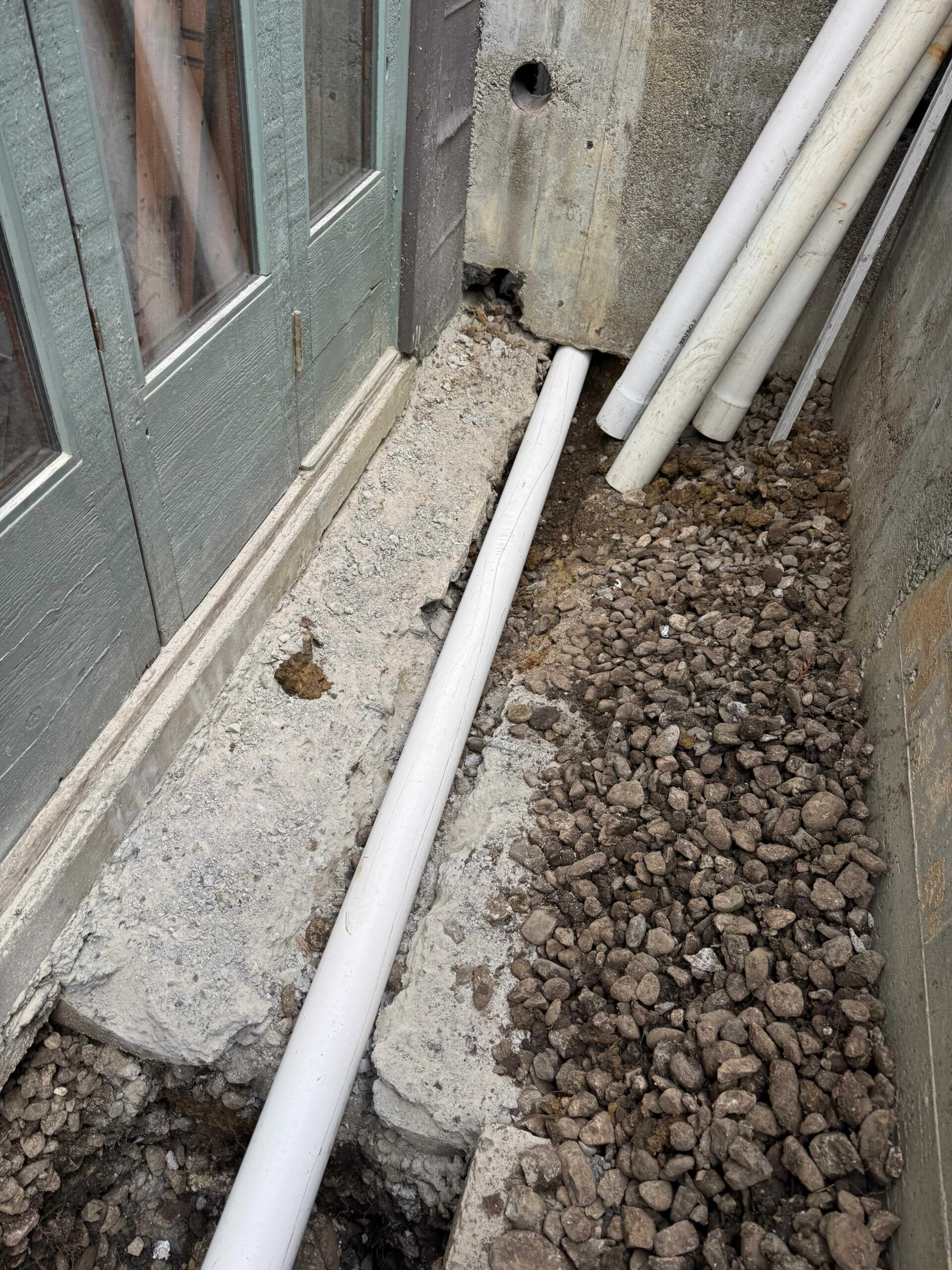 Subsurface Yard Drainage and Foundation Protection White PVC pipe laid down a narrow exterior concrete alleyway surrounded by loose gravel and stone aggregate.